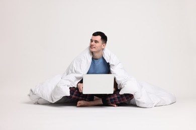 Happy man in pyjama wrapped in blanket using laptop on light grey background