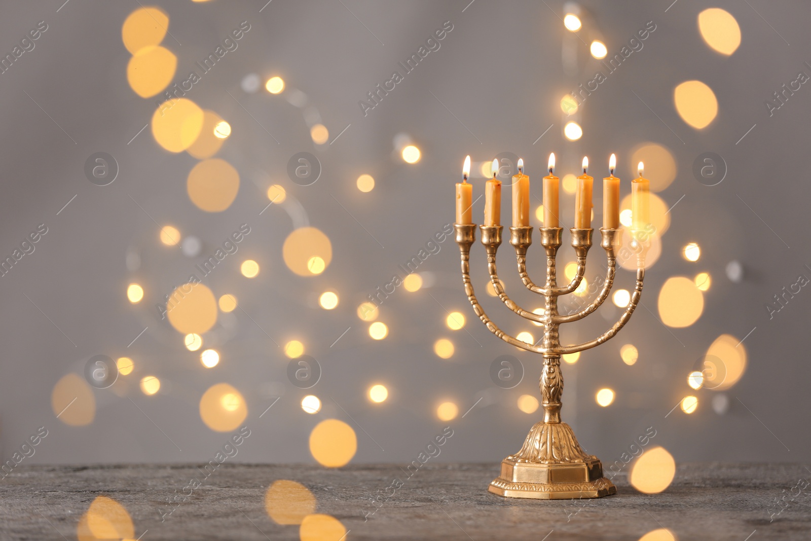 Photo of Golden menorah with burning candles on table against grey background and blurred festive lights, space for text