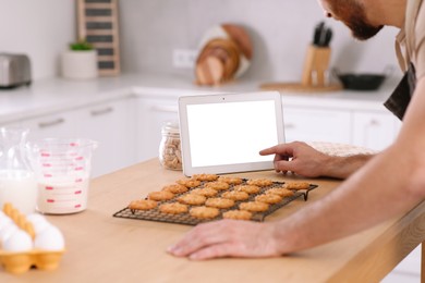 Photo of Man with freshly baked cookies watching online cooking course via tablet in kitchen, closeup