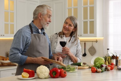 Photo of Happy senior couple cooking together in kitchen. Woman with glass of wine near her husband