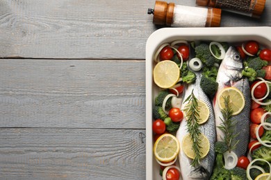 Photo of Raw fish with vegetables and lemon in baking dish and spices on grey wooden table, flat lay. Space for text