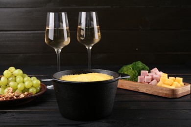 Photo of Fondue pot with melted cheese, glasses of wine and different products on black wooden table