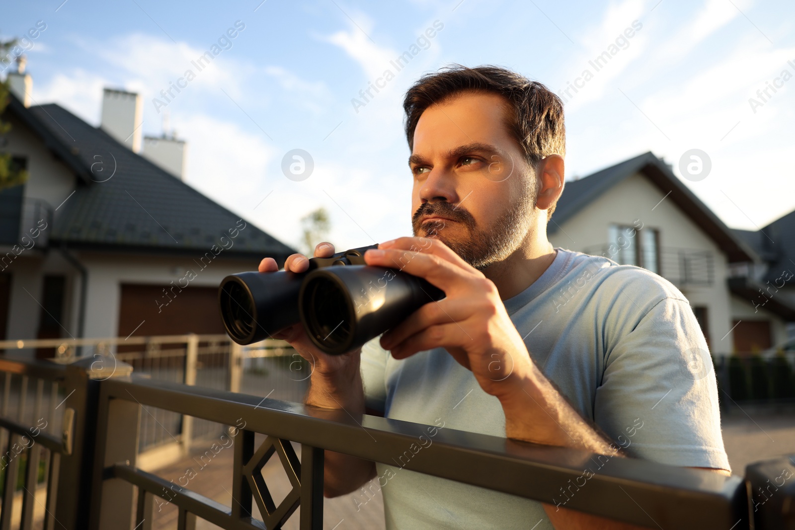 Photo of Concept of private life. Curious man with binoculars spying on neighbours over fence outdoors