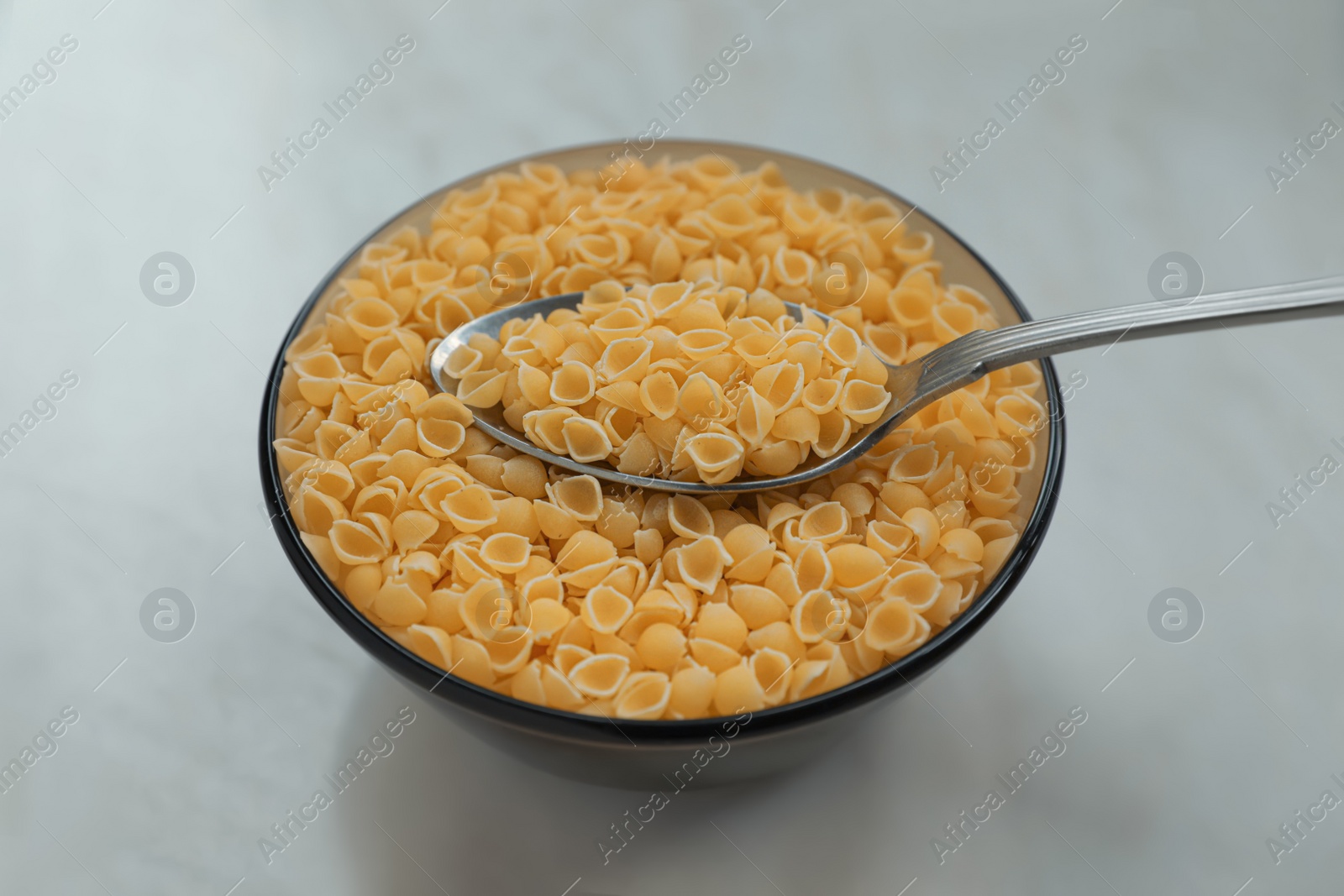 Photo of Bowl of uncooked conchiglie pasta and spoon on white marble table, closeup