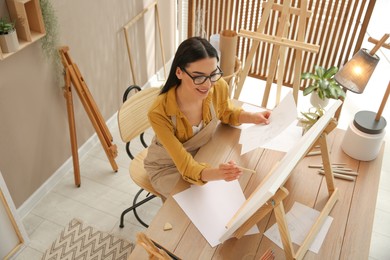 Photo of Young woman drawing on easel with pencil at table indoors, above view