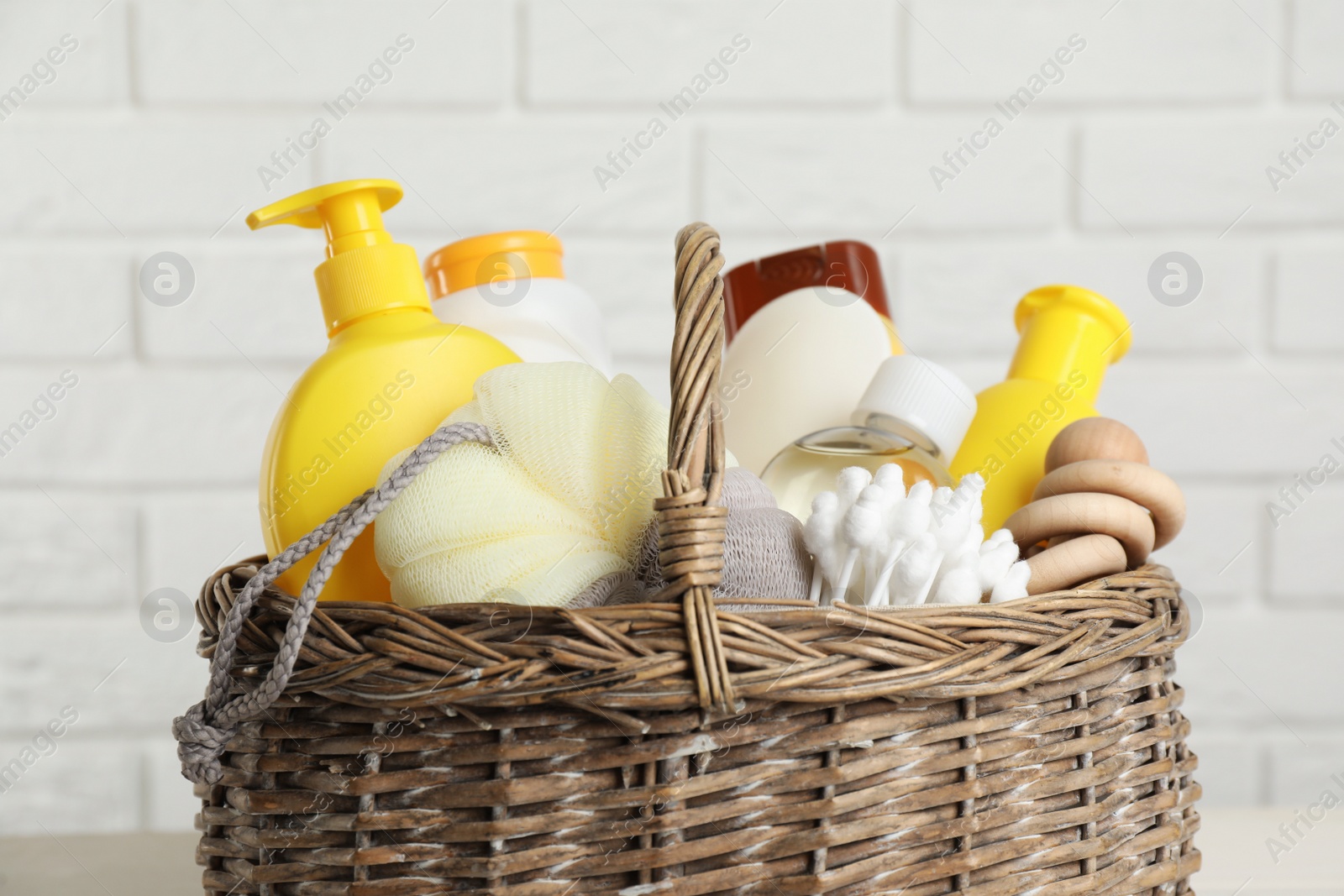 Photo of Wicker basket full of different baby cosmetic products, accessories and toy against white brick wall, closeup