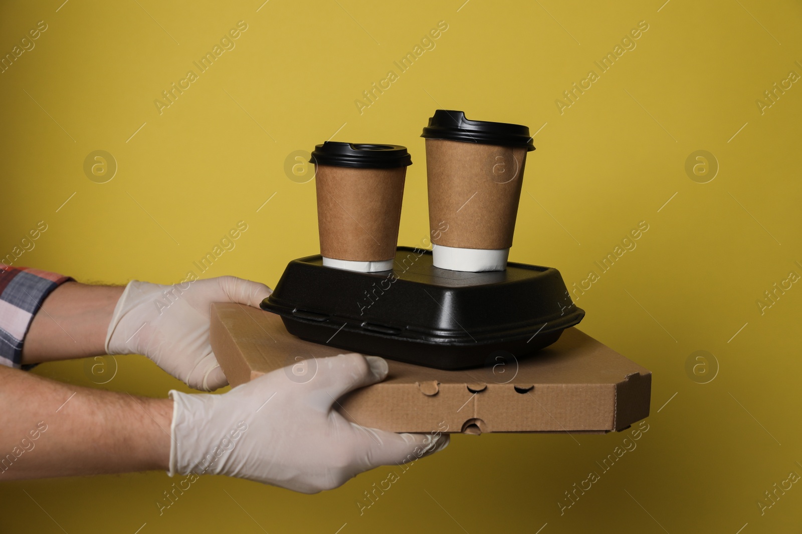 Photo of Courier in protective gloves holding order on yellow background, closeup. Food delivery service during coronavirus quarantine