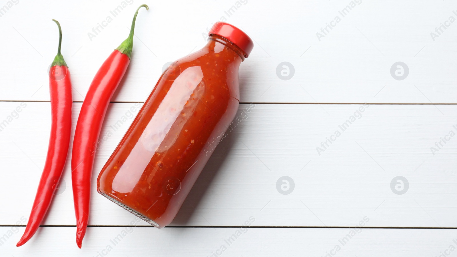 Photo of Spicy chili sauce in bottle and peppers on white wooden table, flat lay. Space for text