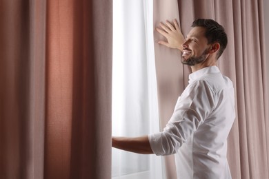 Photo of Happy man opening window curtains at home. Space for text