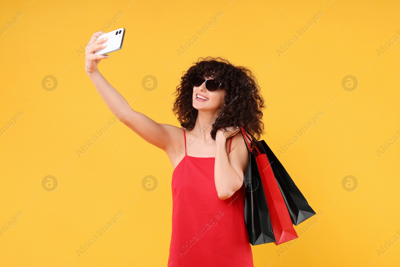 Photo of Happy young woman with shopping bags and stylish sunglasses taking selfie on yellow background