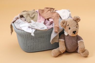 Laundry basket with baby clothes near soft toy on light brown background