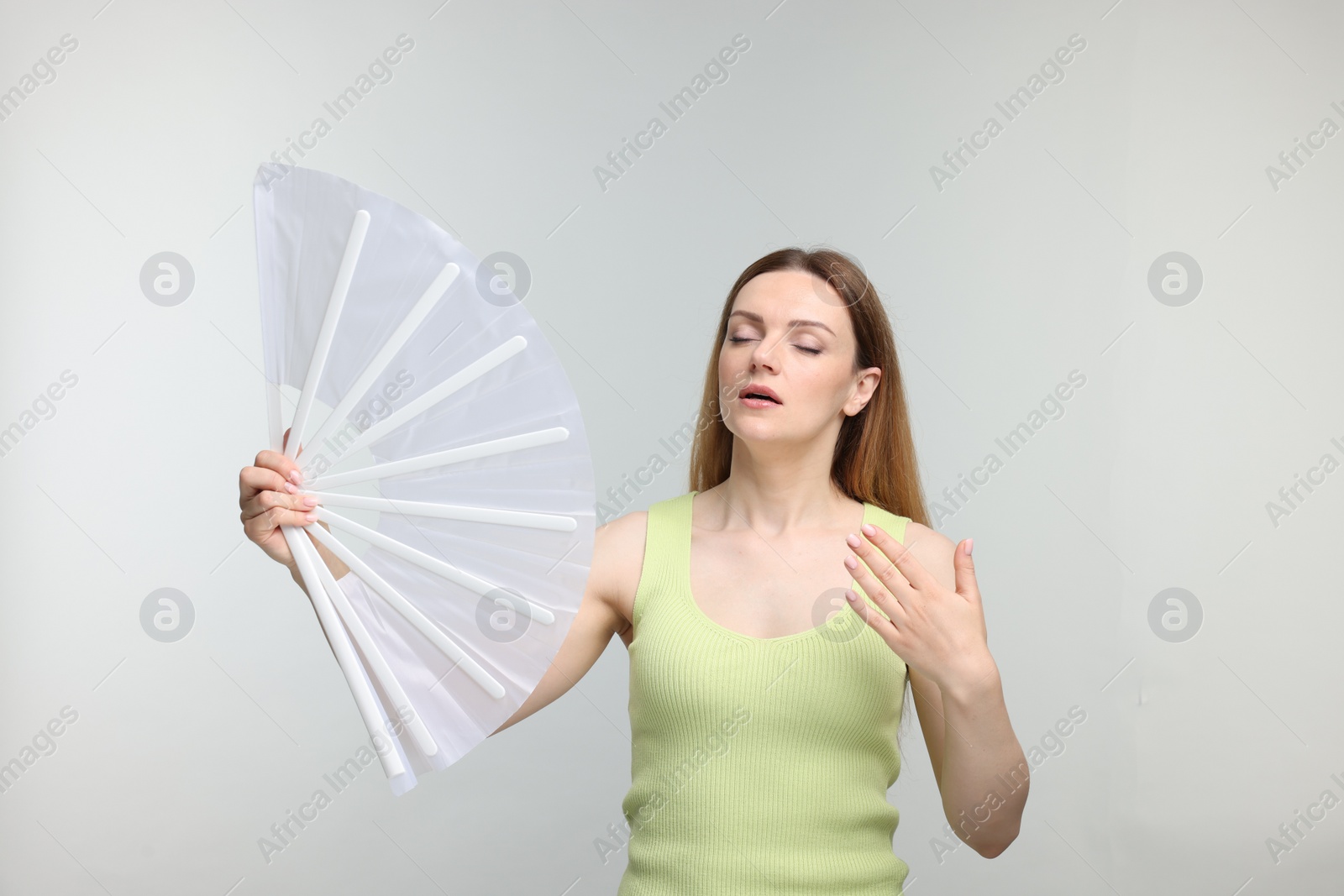 Photo of Beautiful woman waving white hand fan to cool herself on light grey background