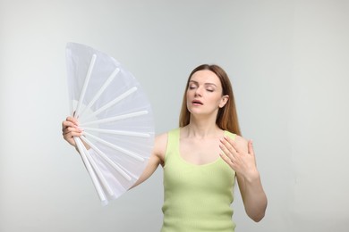 Beautiful woman waving white hand fan to cool herself on light grey background