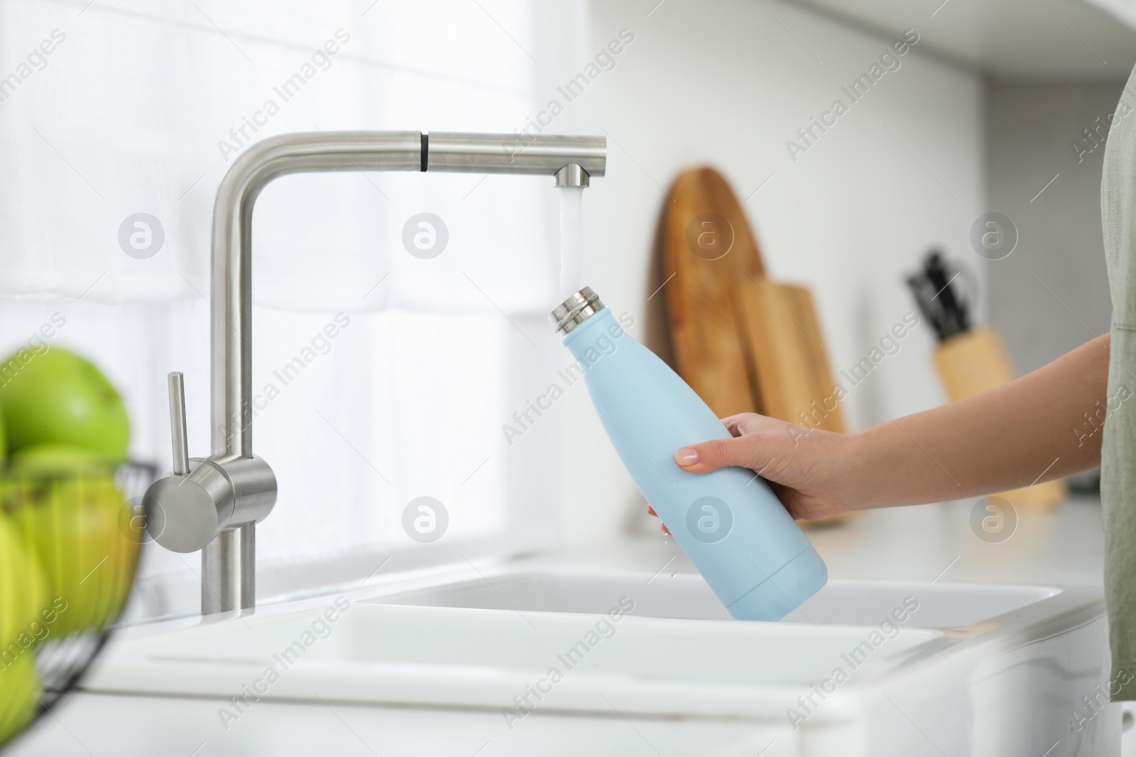 Photo of Woman pouring fresh water from tap into thermo bottle in kitchen, closeup