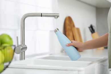 Woman pouring fresh water from tap into thermo bottle in kitchen, closeup