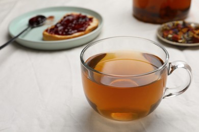 Photo of Cup of freshly brewed tea, dried herbs and bread with jam on white fabric