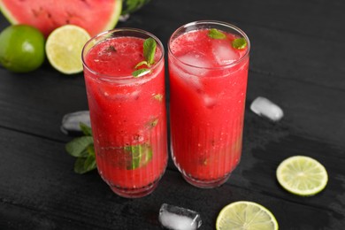 Photo of Glasses of delicious fresh watermelon juice, lime, mint and ice on black wooden table