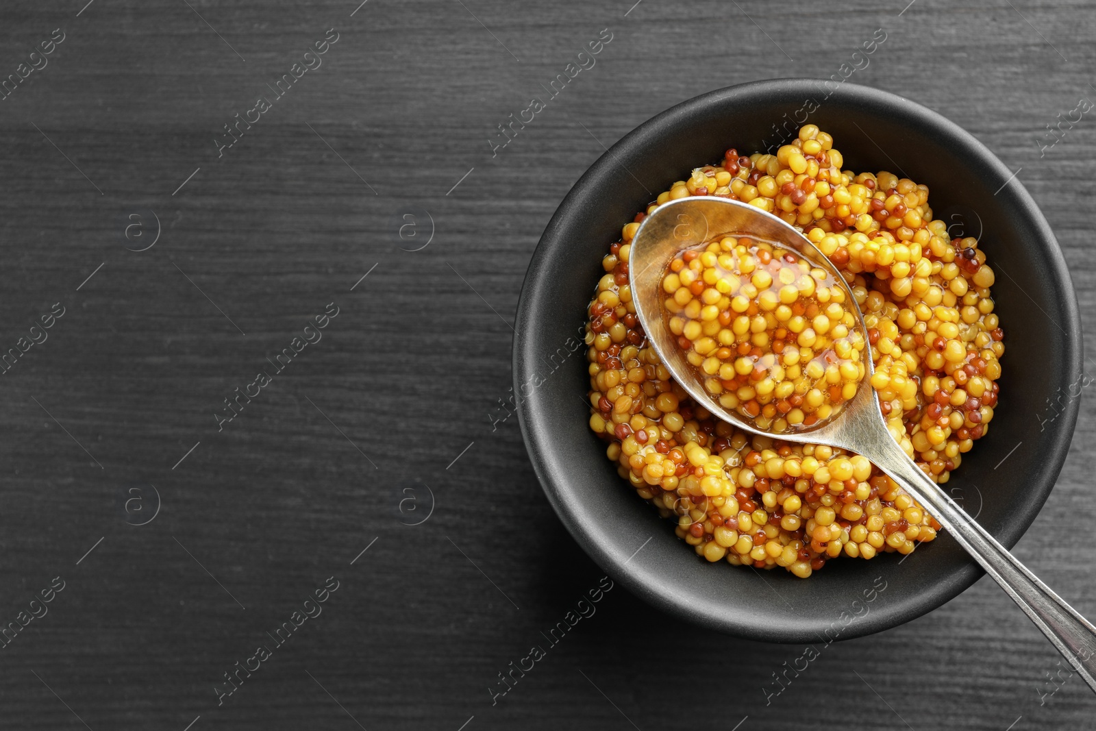 Photo of Whole grain mustard in bowl and spoon on black wooden table, top view. Space for text