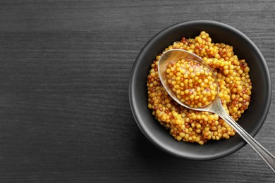 Photo of Whole grain mustard in bowl and spoon on black wooden table, top view. Space for text