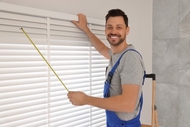 Worker in uniform using measuring tape while installing horizontal window blinds indoors