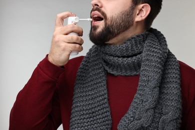 Young man with scarf using throat spray on grey background, closeup