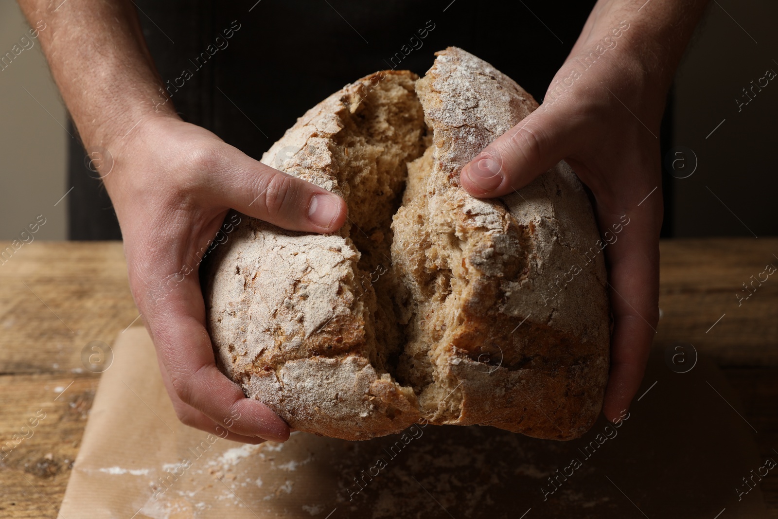 Photo of Man breaking loaf of fresh bread at wooden table, closeup