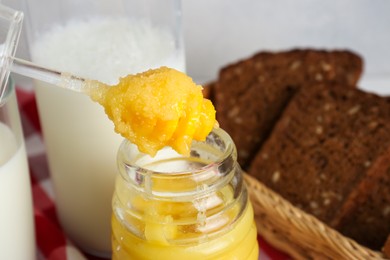 Taking tasty honey from jar with spoon near bread and milk, closeup