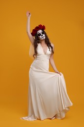 Photo of Young woman in scary bride costume with sugar skull makeup and flower crown posing on orange background. Halloween celebration