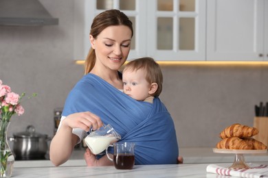 Photo of Mother adding milk into cup of drink while holding her child in sling (baby carrier) in kitchen