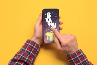 Image of Bonus gaining. Man using smartphone on yellow background, top view. Illustration of open gift box, word and confetti on device screen