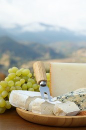 Different types of delicious cheeses, nuts and grapes on wooden table against mountain landscape