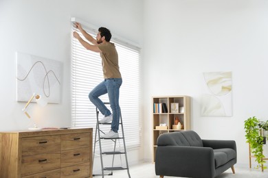 Photo of Man on metal folding ladder installing blinds at home. Space for text