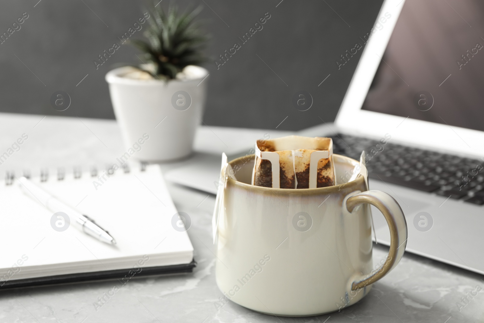 Photo of Cup with drip coffee bag, laptop and notebook on light grey marble table, closeup
