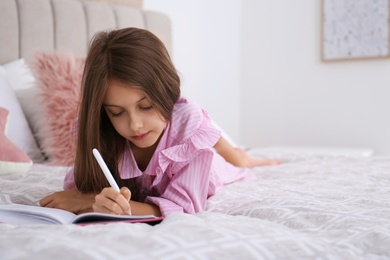 Cute little girl writing in notebook on bed at home