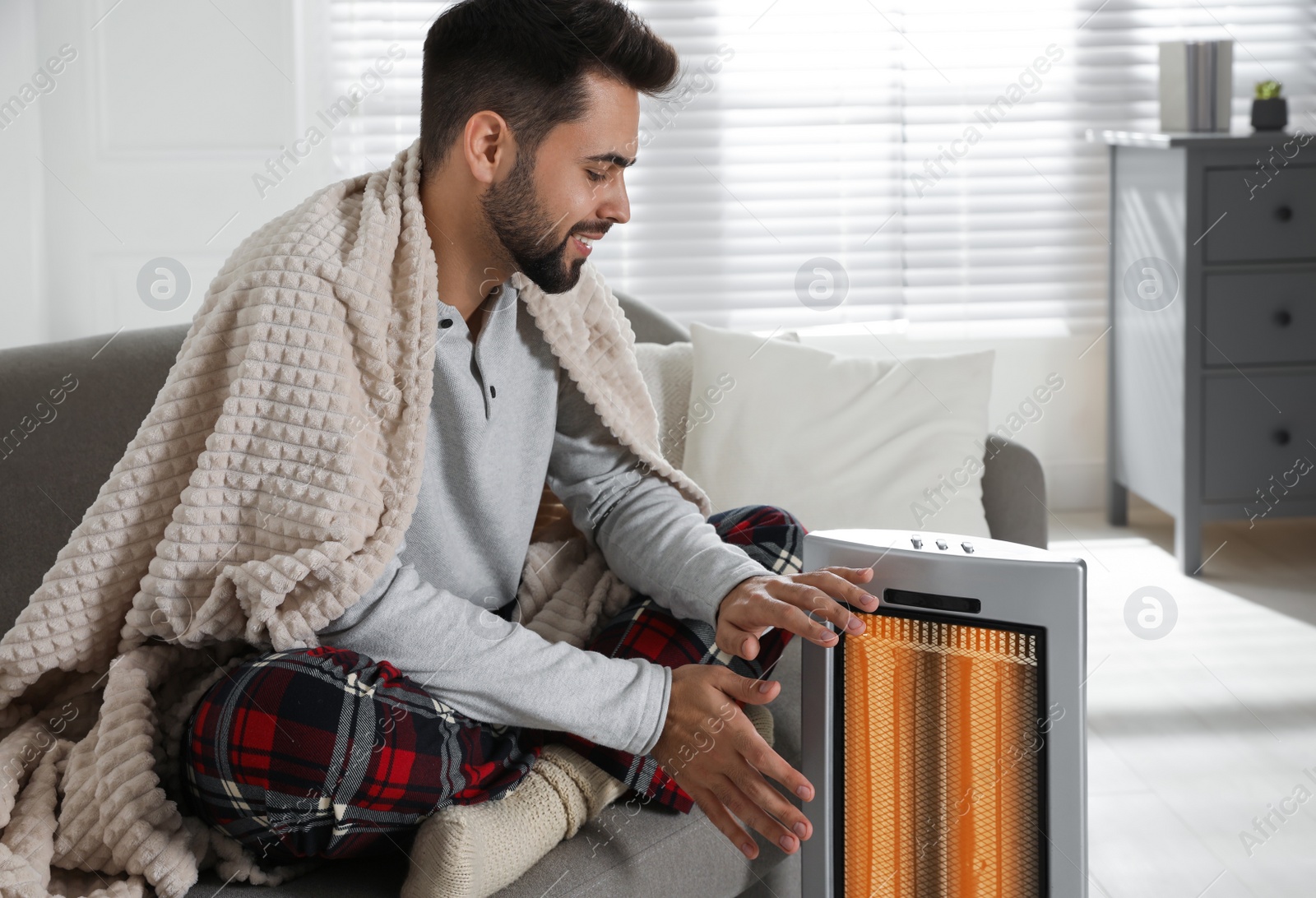 Photo of Young man warming hands near electric heater at home