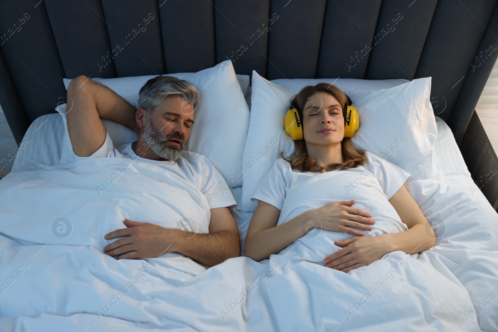 Photo of Smiling woman with headphones lying near her snoring husband in bed at home