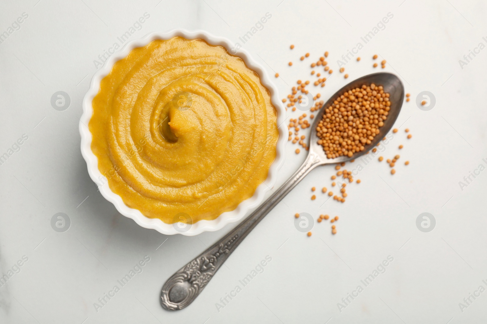 Photo of Tasty mustard sauce in bowl and spoon with dry seeds on white table, flat lay