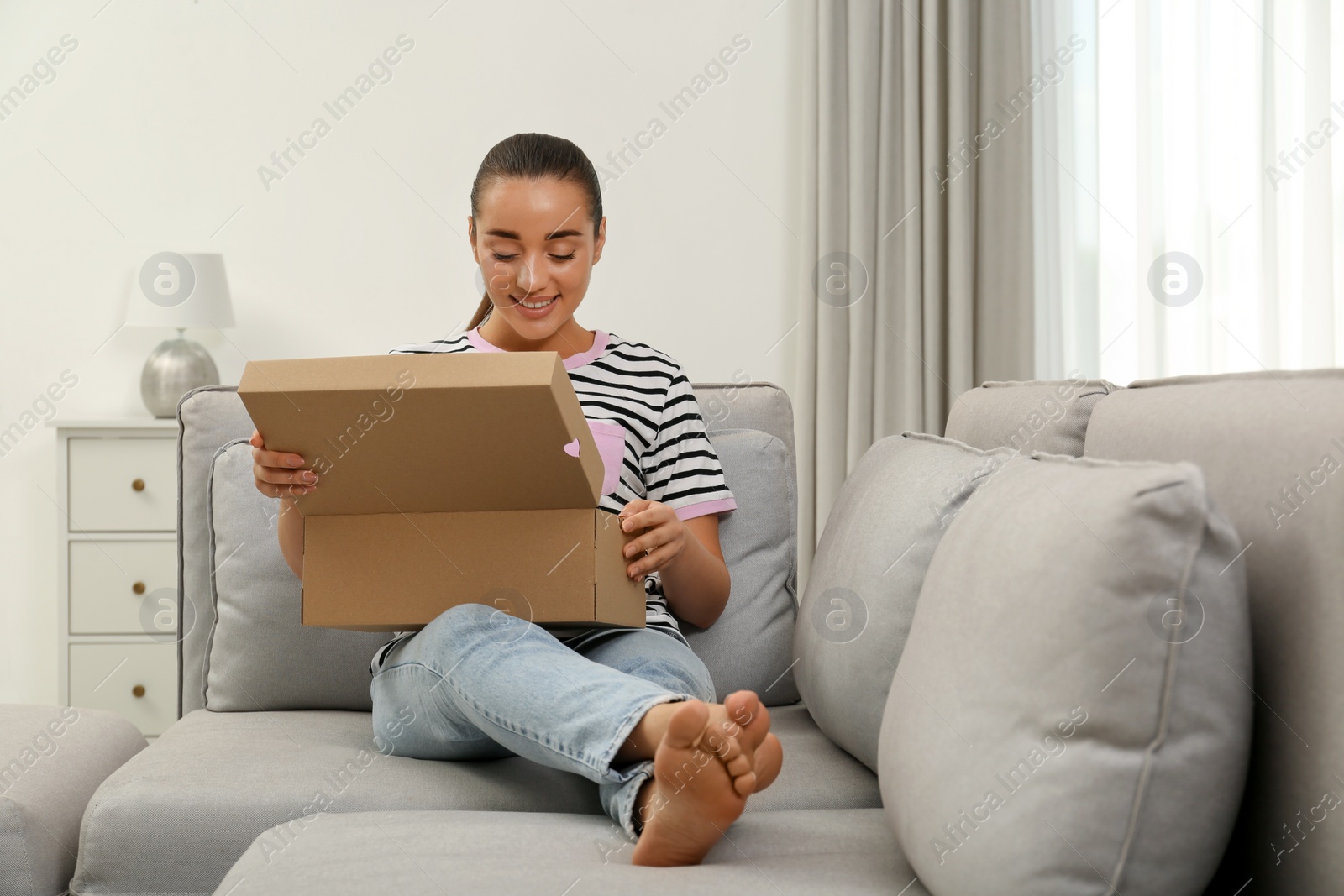 Photo of Happy young woman opening parcel on sofa at home. Internet shopping
