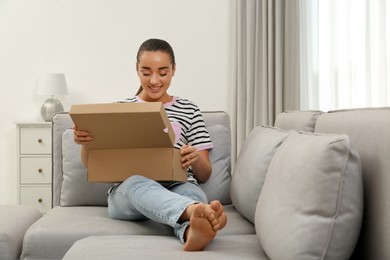 Happy young woman opening parcel on sofa at home. Internet shopping