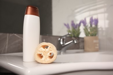 Photo of Natural loofah sponge and bottle of shower gel on washbasin in bathroom, space for text