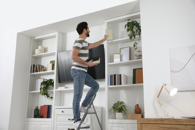 Photo of Man on metal folding ladder taking book from shelf at home