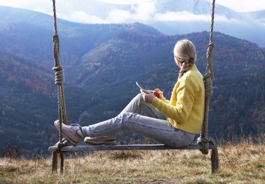 Photo of Young woman drawing with graphic tablet while sitting on swing in mountains