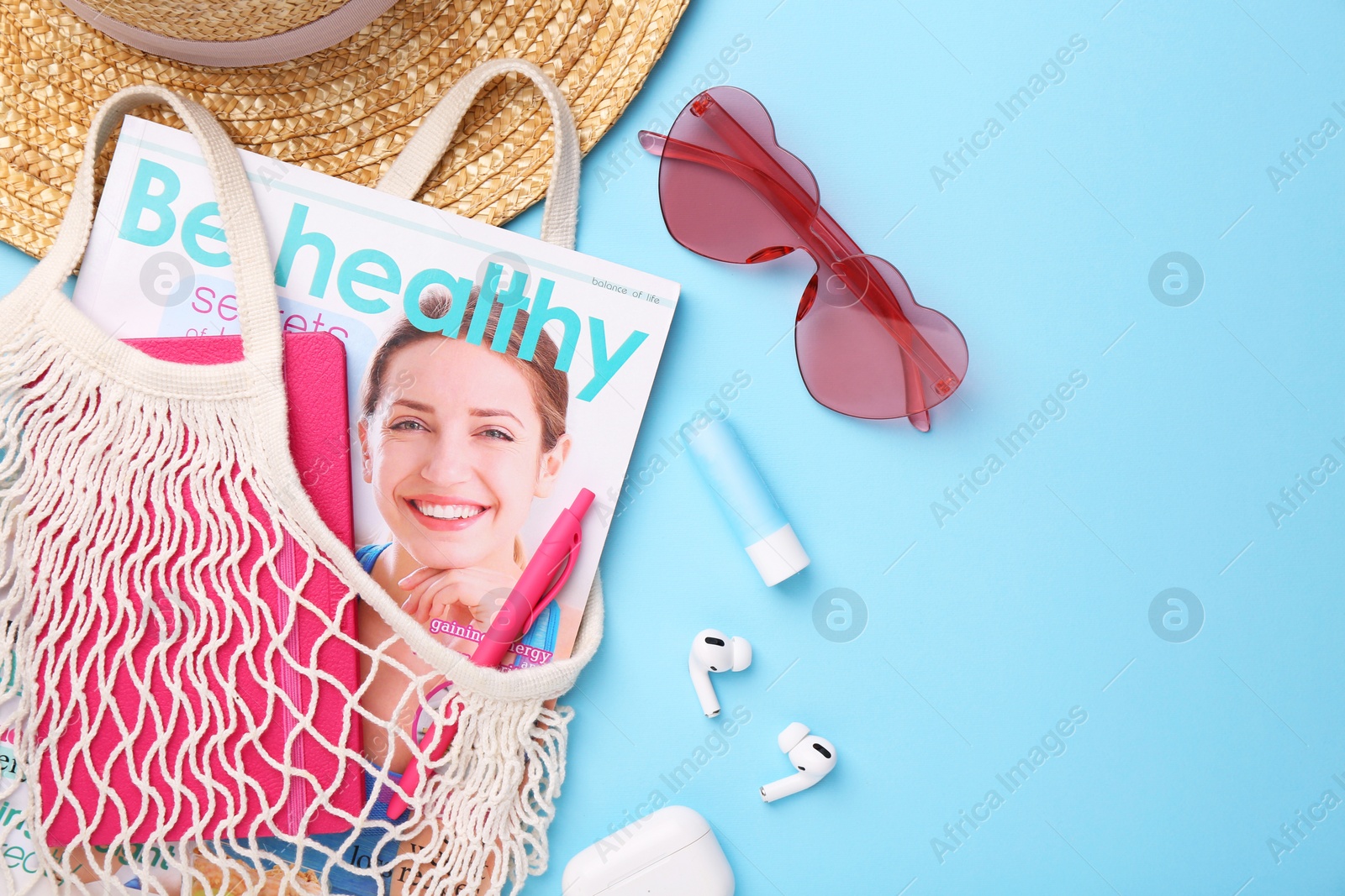 Photo of String bag with magazine, earphones and summer accessories on light blue background, flat lay. Space for text