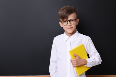 Photo of Cute schoolboy in glasses with book near chalkboard, space for text