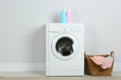 Modern washing machine with detergents and laundry basket near white wall