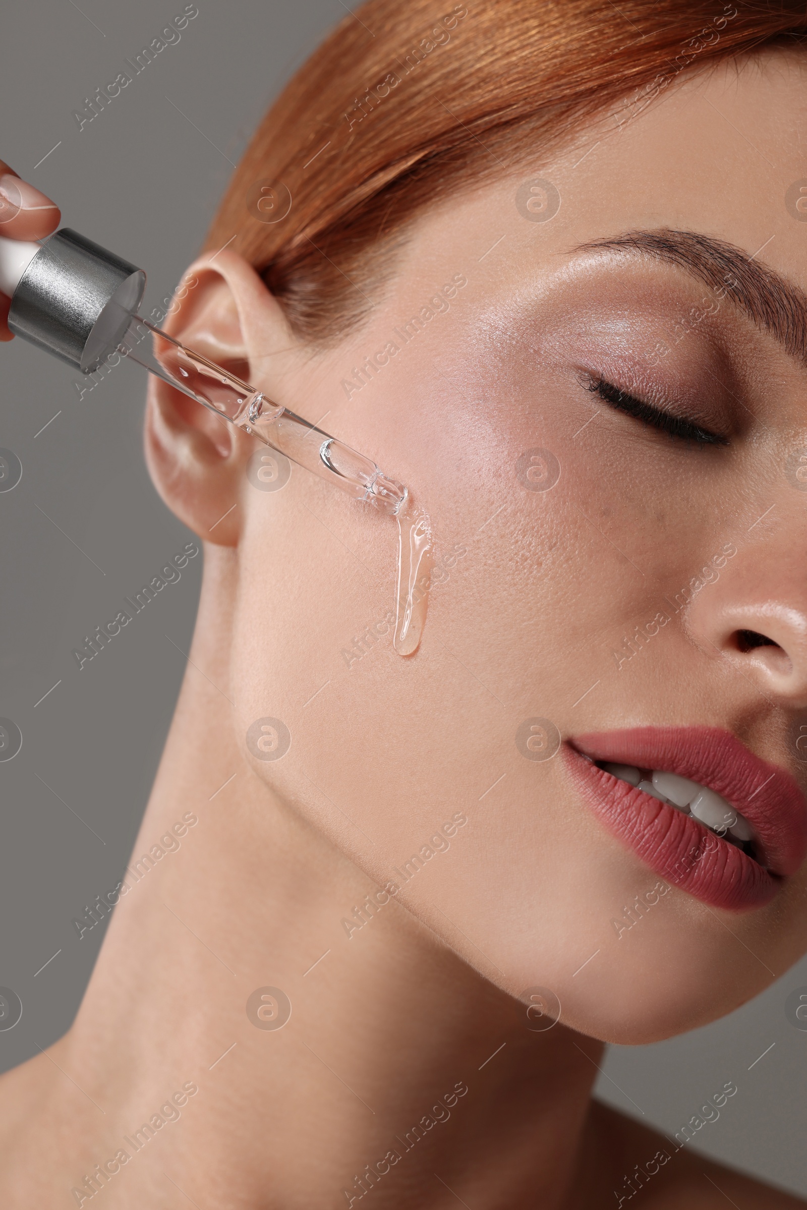 Photo of Beautiful young woman applying cosmetic serum onto her face on grey background, closeup