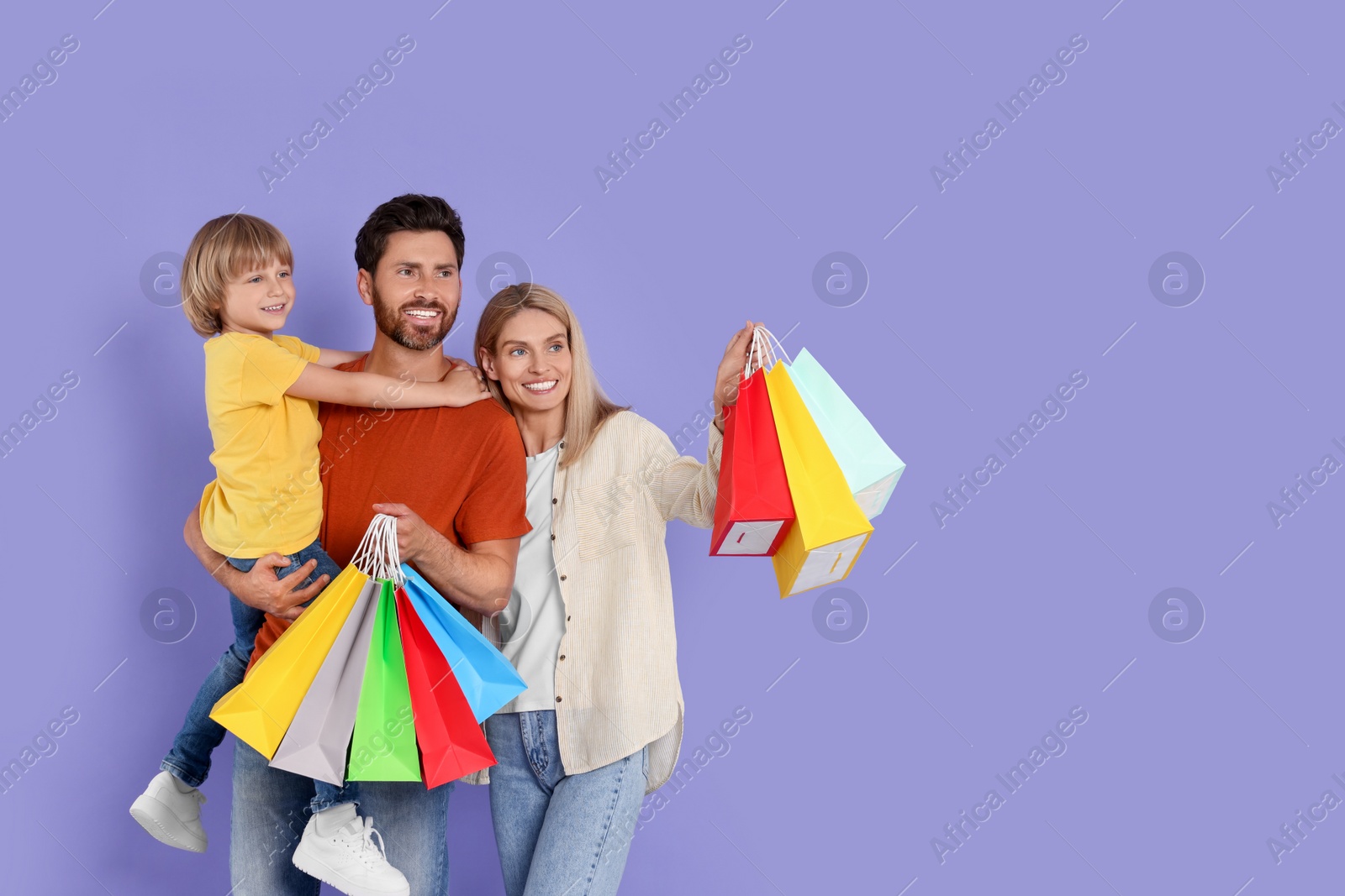 Photo of Family shopping. Happy parents and son with many colorful bags on violet background. Space for text