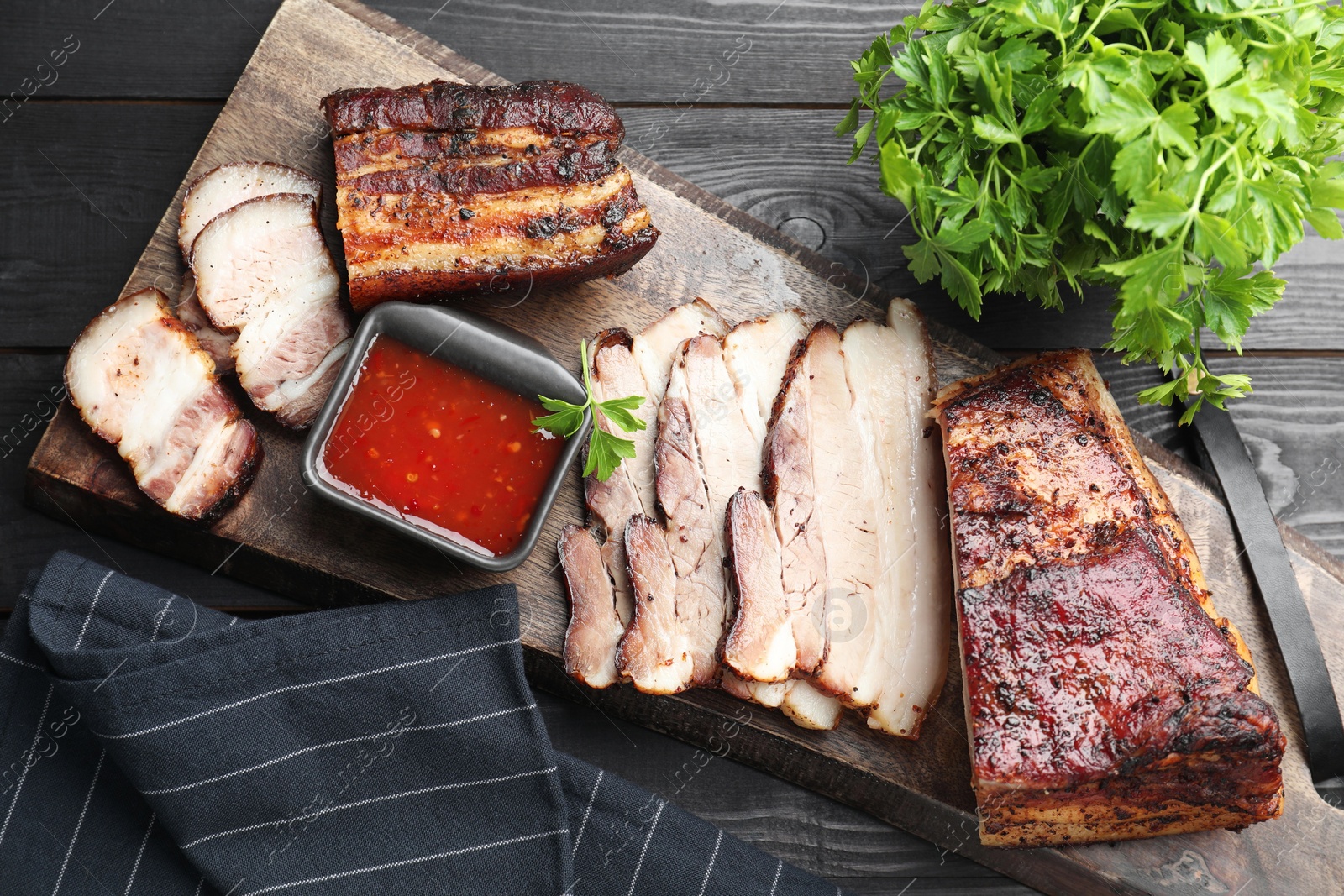 Photo of Pieces of baked pork belly served with sauce and parsley on black wooden table, top view