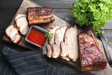 Photo of Pieces of baked pork belly served with sauce and parsley on black wooden table, top view