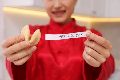 Photo of Young woman holding tasty fortune cookie with prediction Yes, You Can in kitchen, closeup